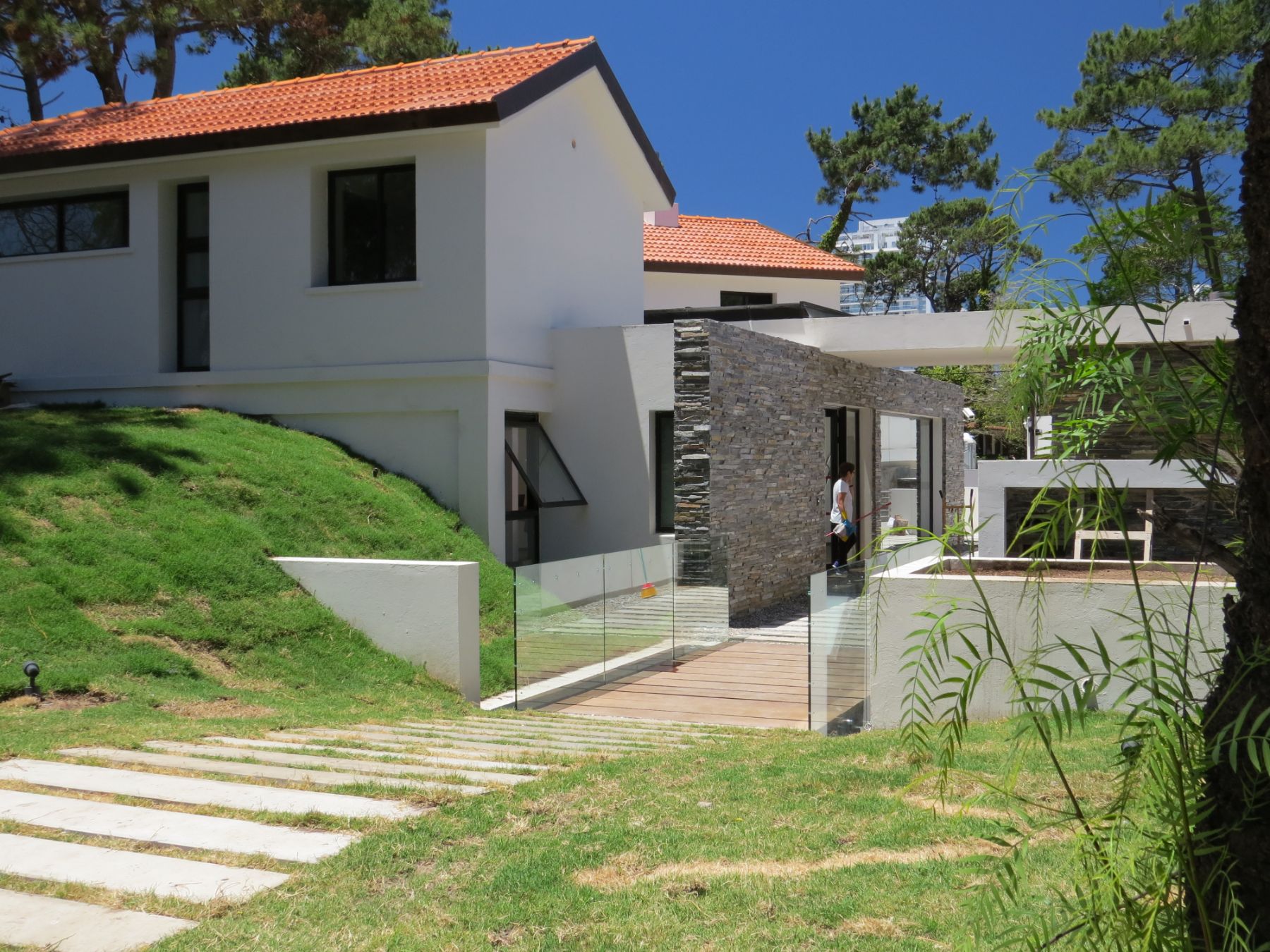 En la imagen se aprecia la entrada principal de la policlínica de punta del este, en la que se puede ver un puente de madera, una baranda de vidrio, un jardín, una pared revestida de piedra, dos tejados a dos aguas cada uno.